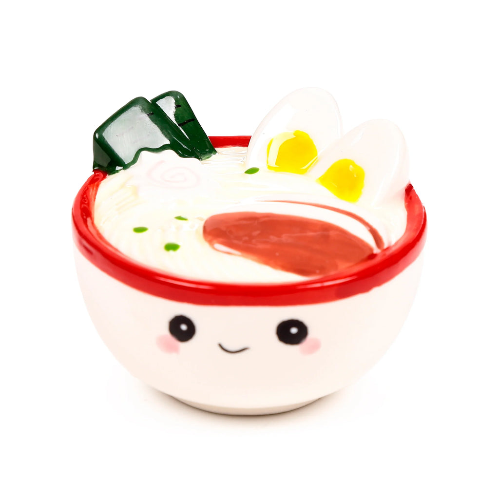 Front view of a white ceramic ramen bowl shaker with a red rim and smiling face, featuring imitation ramen toppings: two white egg halves with yellow centres, a pink swirl, green seaweed pieces, and a red oval shape.