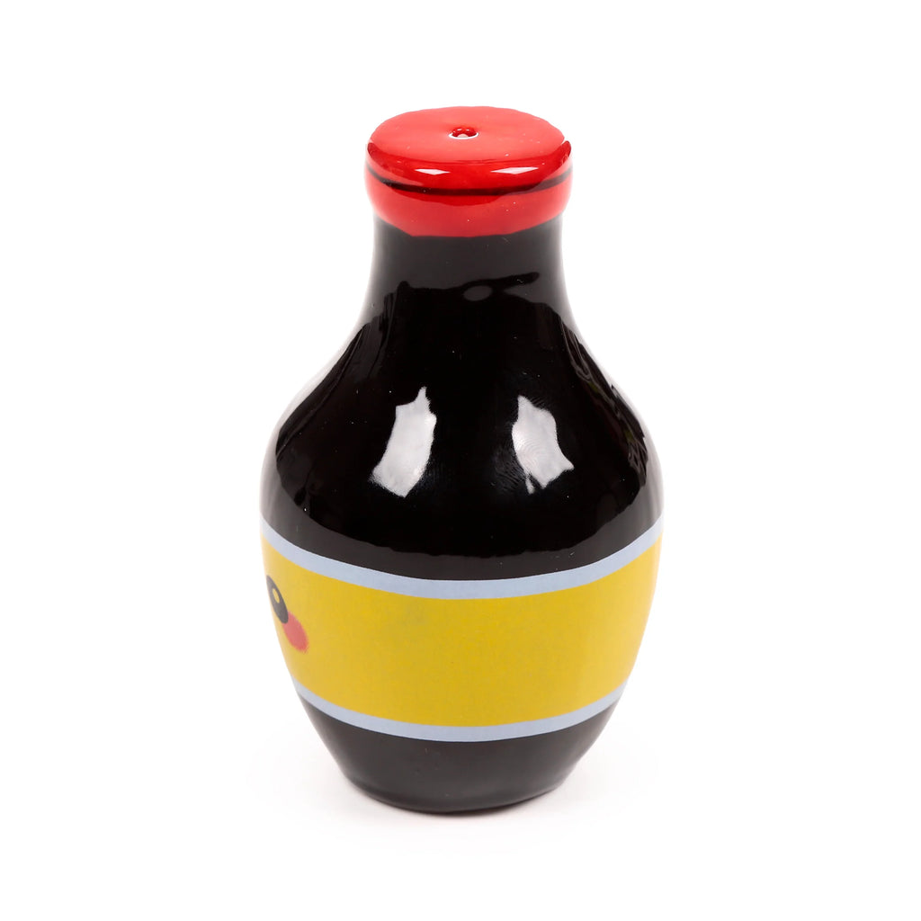 Front view of a black ceramic salt or pepper shaker, shaped like a ramen sauce bottle, with a red cap, a yellow central band, and a partially visible cartoon Foodiemal face.