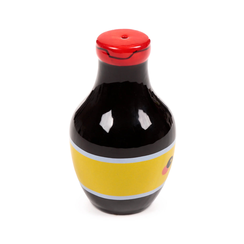 Angled view of a black, red, and yellow ceramic salt or pepper shaker, shaped like a stylised soy sauce bottle, with a red lid featuring a single hole and a yellow band depicting a character's eye and cheek.