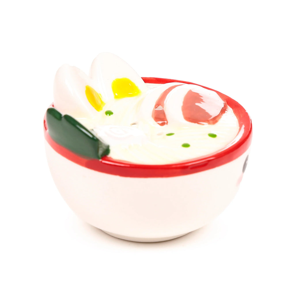 Angled view of a ceramic salt and pepper shaker shaped like a ramen bowl, with a white body, red rim, a small black eye and pink blush on the side, showing white noodles, two egg halves with yellow yolks, a red swirl, and green garnishes.