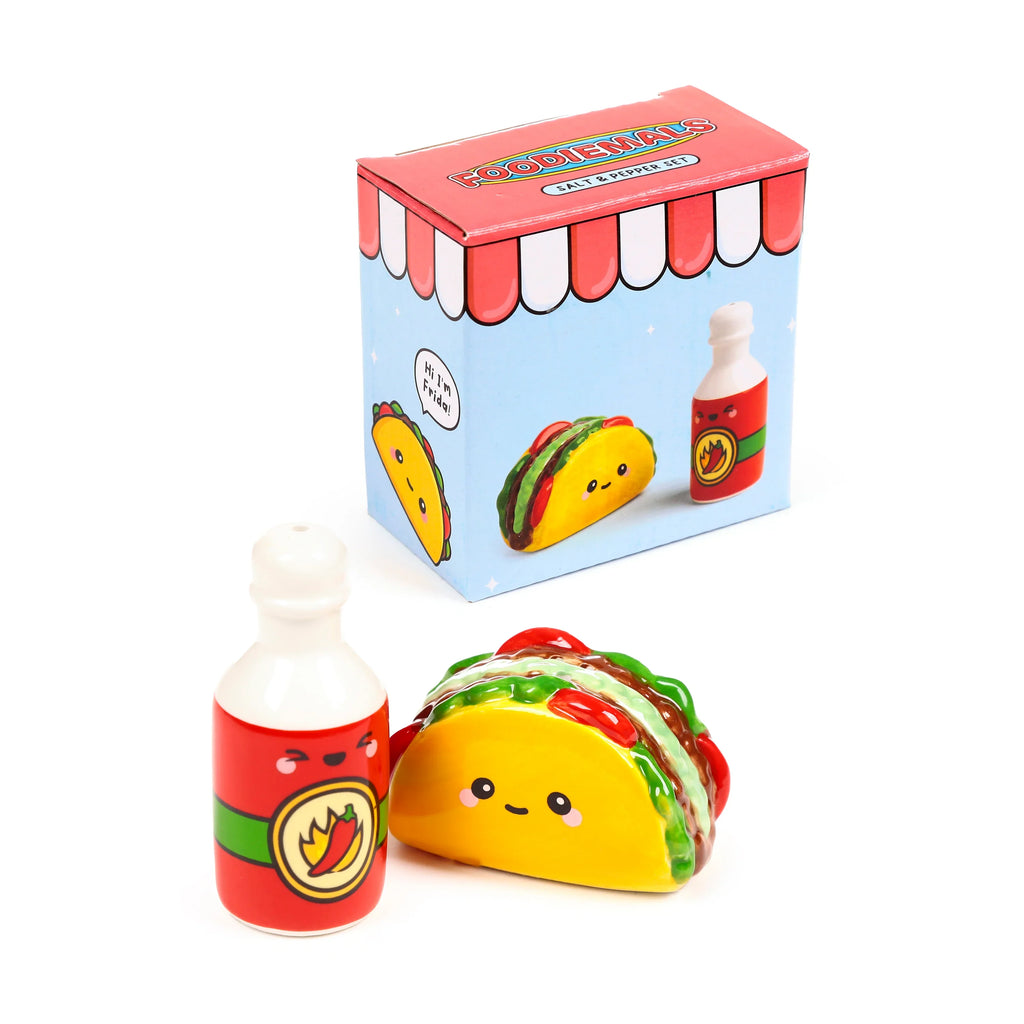 Angled studio shot of a ceramic taco salt shaker and matching hot sauce bottle pepper shaker, both with cheerful faces, presented with their colourful, illustrated packaging on a white background.