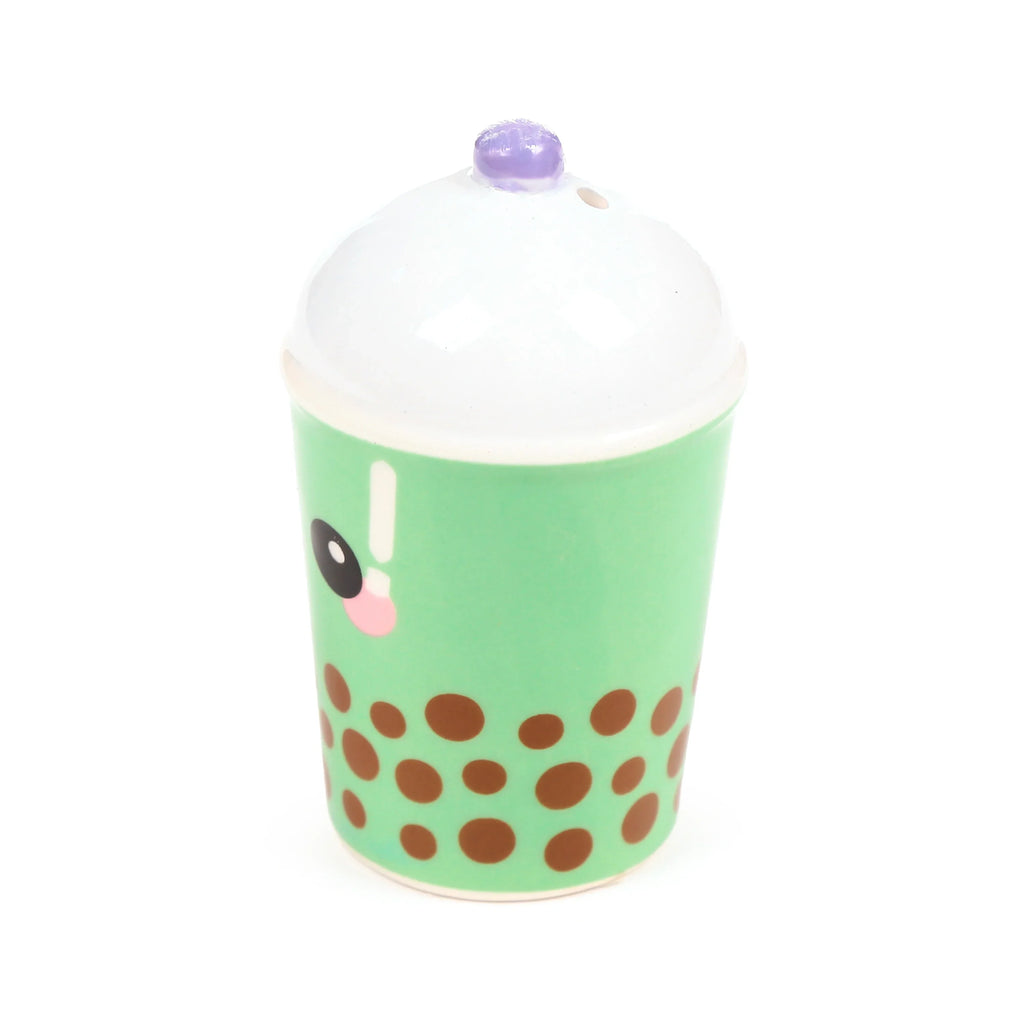 Angled front view of a ceramic shaker shaped like a light green matcha bubble tea cup, featuring brown boba pearls, a cartoon face with a black eye and pink blush, a white foam-like top, and a purple straw-like detail.