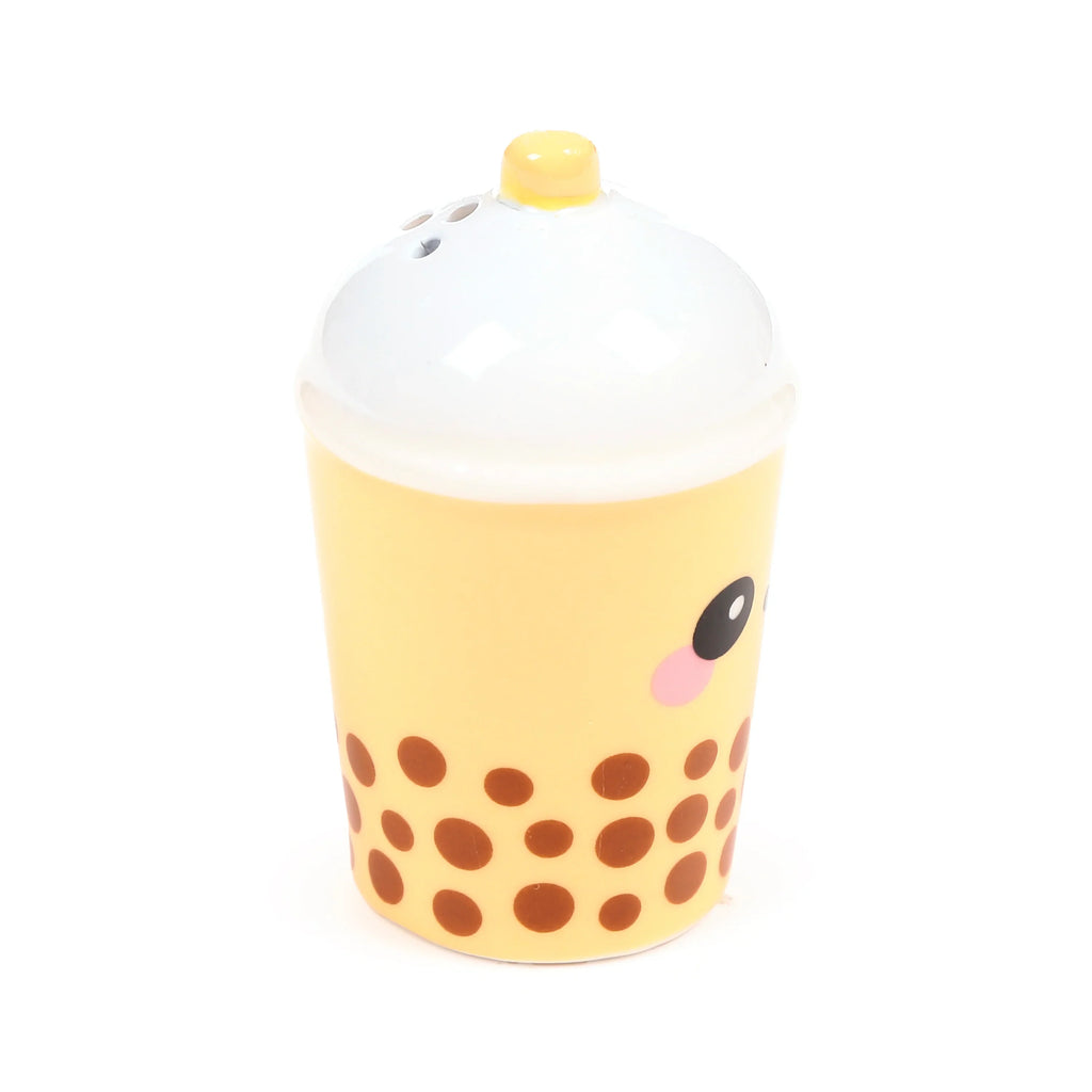 Front view of a light yellow ceramic salt shaker, shaped like a boba tea cup with brown tapioca pearl details. It features a cartoon face with a black eye and pink blush, topped with a white domed lid and small yellow straw cap, showing shaker holes.