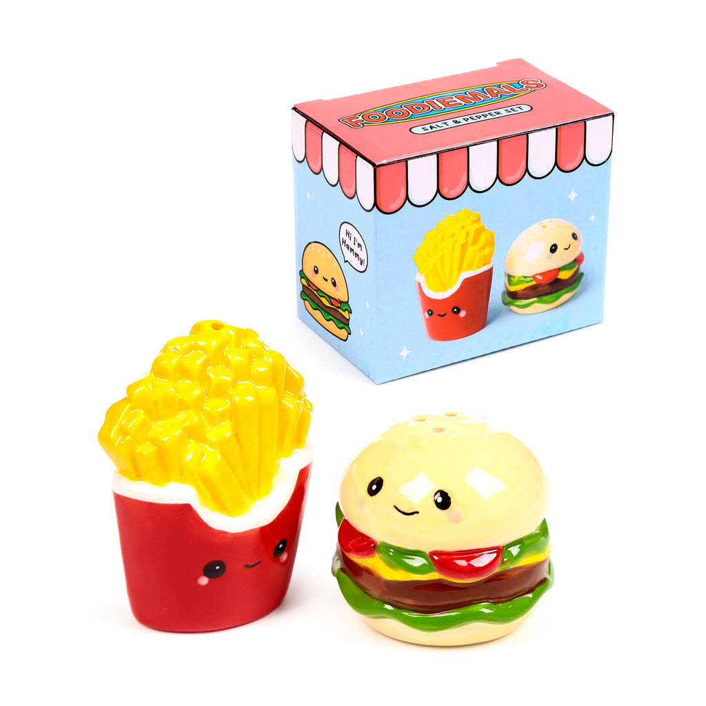 Angled view of a ceramic burger and fries salt and pepper set. The smiling burger and fries shakers, both with shaker holes, are placed in front of their colourful Foodiemals box, which features cartoon illustrations and a red and white striped top.