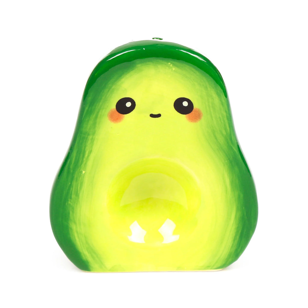 Front view of a ceramic avocado shaker, shiny and green, with a cartoon face, black eyes, small smile, and pink cheeks. It features a circular indentation in its lighter green centre, set against a white background.
