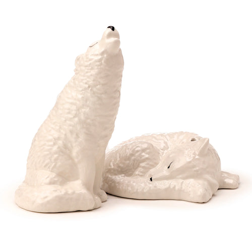 A pair of white ceramic novelty wolf salt and pepper shakers, one sitting with head raised and the other curled up sleeping, on a white background.