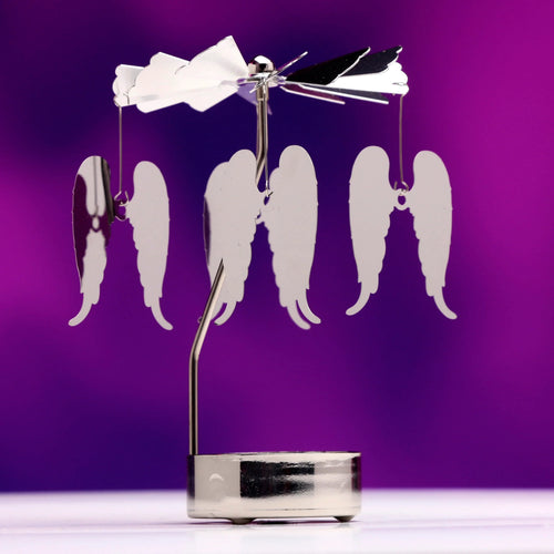 A straight-on view of a silver-coloured spinning tea light carousel candle holder, featuring four suspended angel wing decorations and a shiny metallic finish, set against a purple background.