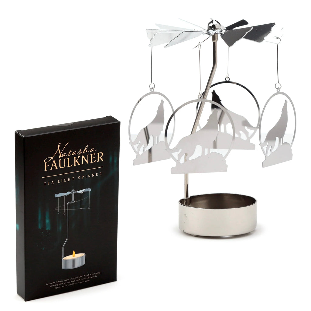 Angled view of a metallic silver tea light carousel candle holder, featuring four dangling howling wolf silhouette charms and a rotating vane top. A black product packaging box is visible to the left.