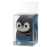 Front view of a dark grey and white penguin makeup sponge, with black eyes, an orange beak, pink cheek blushes, and a 'BRRR' speech bubble, packaged in a clear plastic box featuring blue ice-like graphics and 'MAKEUP SPONGE' text.