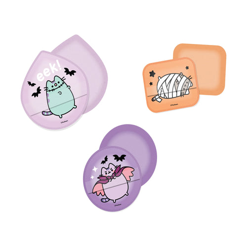 Overhead view of three Halloween Pusheen the Cat beauty blender sponges. A purple tear-drop sponge features a Frankenstein Pusheen, an orange rectangular sponge shows a mummy Pusheen, and a purple circular sponge depicts a vampire Pusheen.