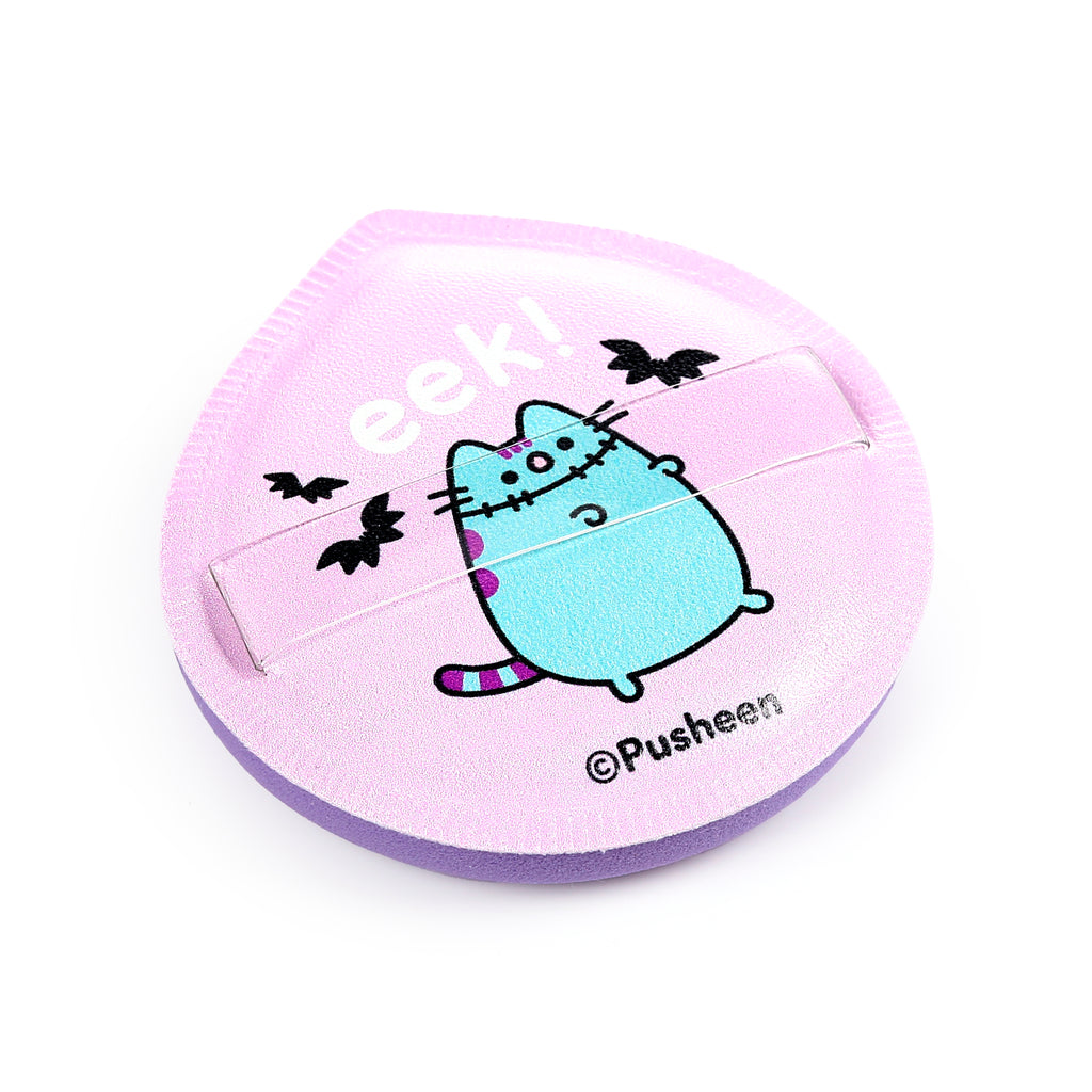 Angled top view of a light purple flat makeup sponge pad, featuring a Halloween-themed blue Pusheen cat with stitched details, 'eek!' text, and black bat illustrations, with a clear finger strap across its surface.