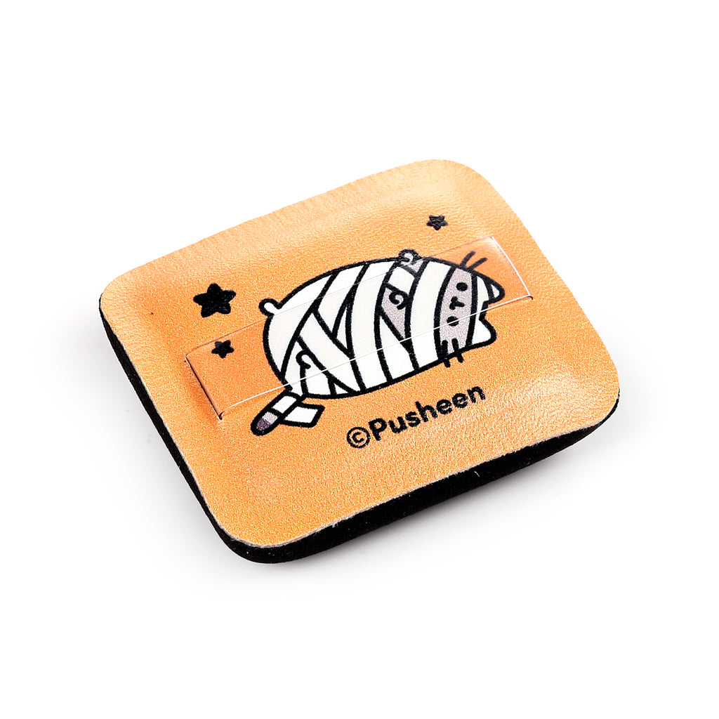 Angled view of a square, textured orange makeup accessory compact, depicting a mummy Pusheen cat design with clear film detail, black star motifs, and dark edges.