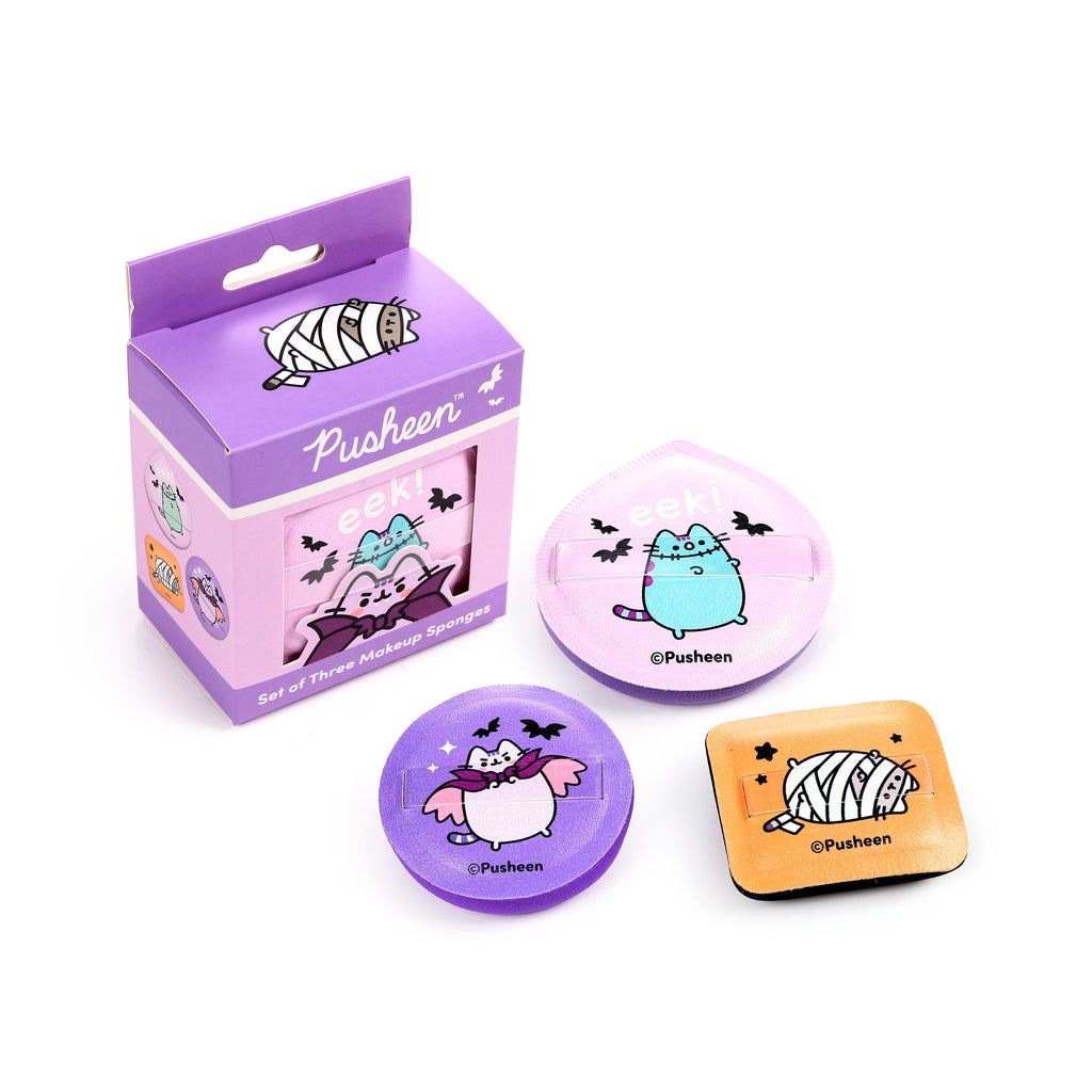 Overhead angled view of a purple box containing three Halloween Pusheen beauty sponges, displayed alongside the individual sponges. The set includes a pink sponge with a blue Frankenstein cat, a purple sponge with a white vampire cat, and an orange sponge with a mummy cat design, each featuring a finger strap.
