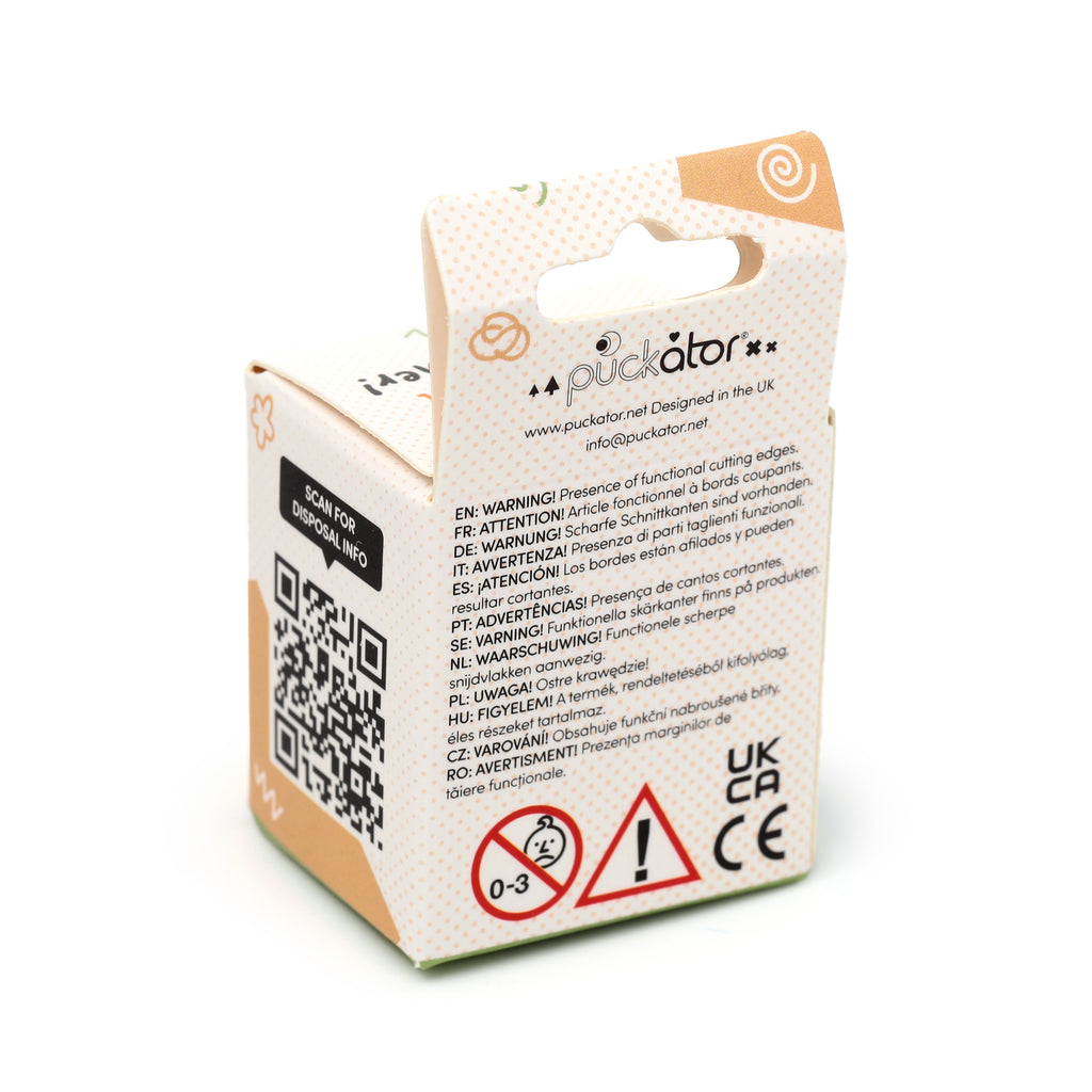 Angled view of the reverse of light beige cardboard packaging for a pencil sharpener and eraser set, showing multilingual warning text about sharp edges, a QR code for disposal, 0-3 age restriction, UKCA, and CE certification marks. The box has a peach polka dot pattern and a top hang tab.