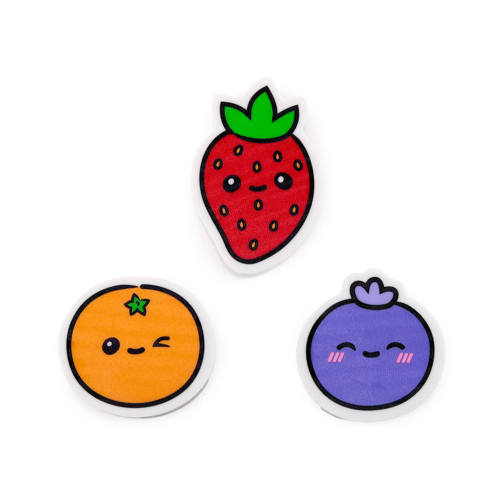 Overhead view of three colourful fruit-shaped erasers: a red strawberry, an orange, and a purple blueberry, each with a cute face, on a white background.
