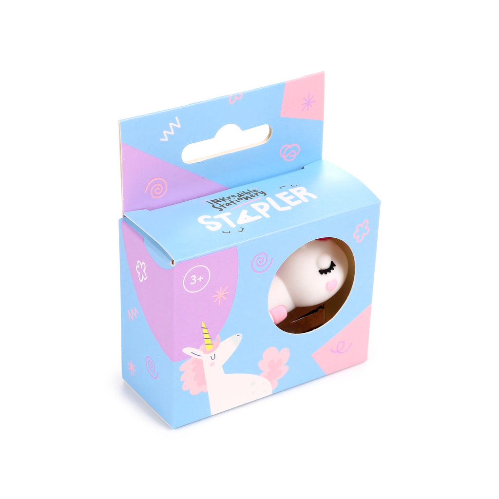 Angled view of a mini unicorn-shaped stapler within its colourful light blue and pink retail box. The packaging features a circular cut-out revealing part of the white unicorn stapler, showing its closed eyes and pink cheeks. The box also displays a unicorn graphic and the word 'Stapler'.