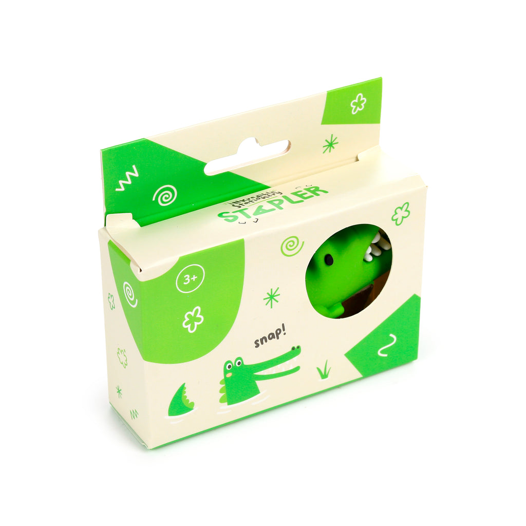 Angled view of a mini crocodile stapler in its cream and green retail packaging. The box displays green cartoon crocodile graphics, abstract patterns, and a circular window showing the stapler's green head and white teeth.