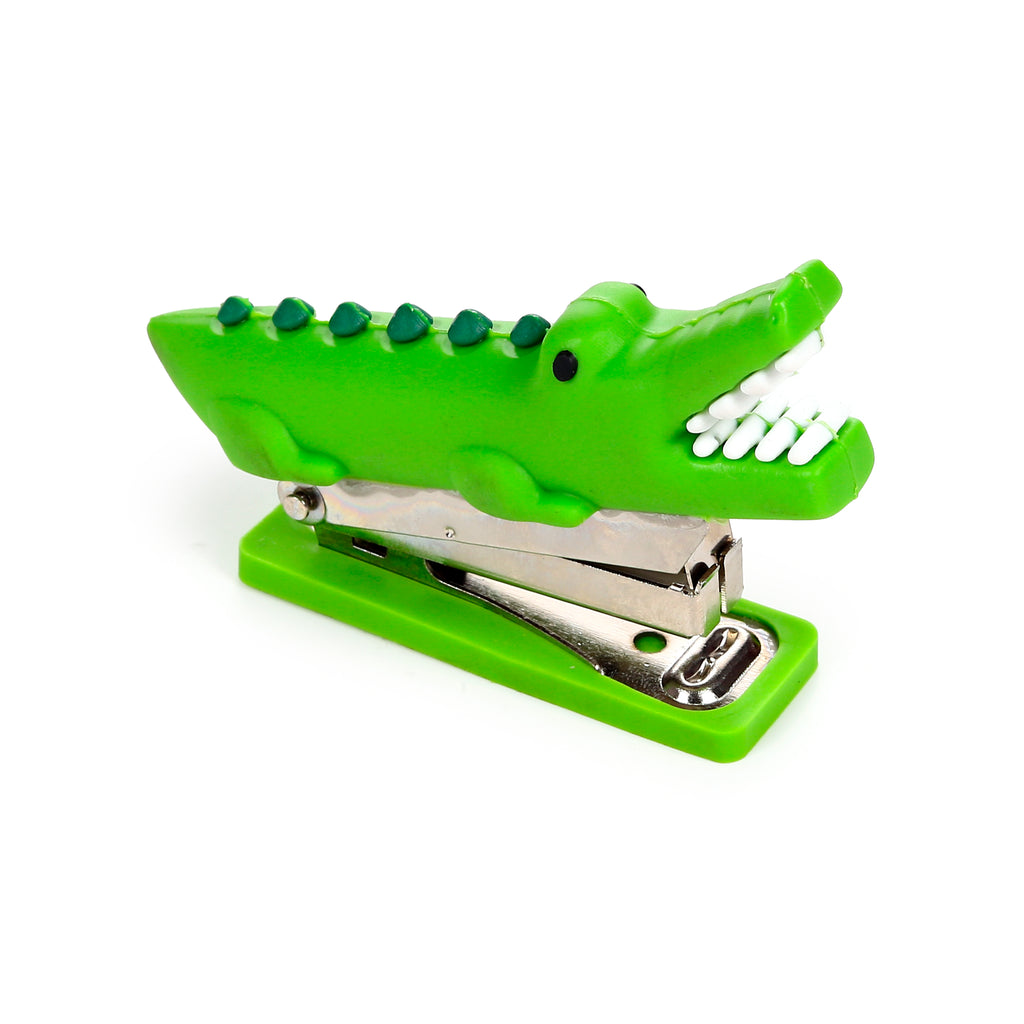 Angled view of a bright green crocodile mini stapler, featuring an open mouth with white teeth, black eyes, and darker green bumps along its back, sitting on a matching green base.