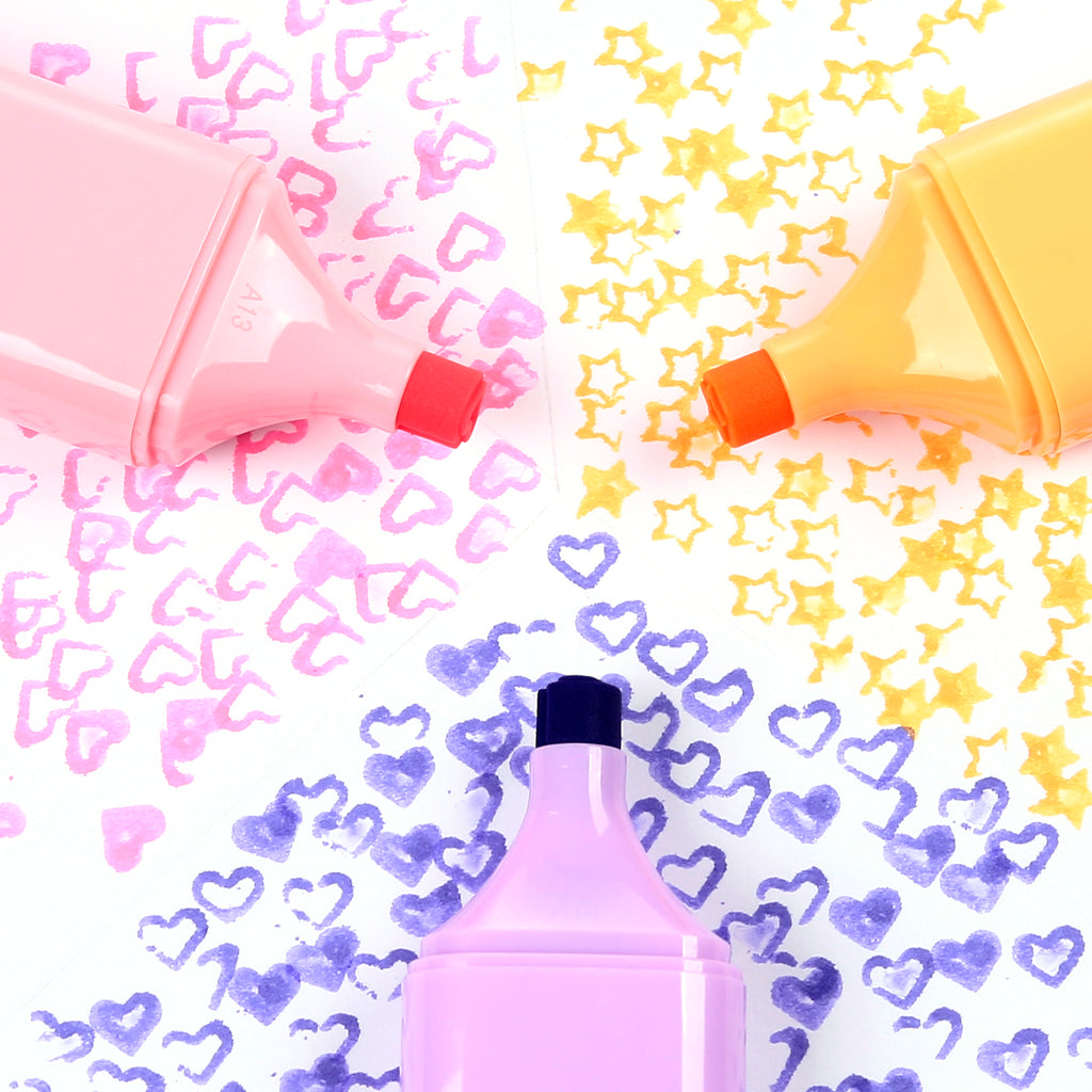 Overhead view of three highlighters in pastel pink, yellow, and lilac, creating pink hearts, yellow stars, and purple heart patterns on a white surface.