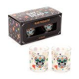 Angled view of two clear glass tumblers, each decorated with a vibrant Day of the Dead sugar skull and colourful patterns. Behind them is their black presentation box, featuring a clear window displaying the tumblers and text 'GLASS TUMBLER SET 2 x 250ml'.