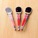 Overhead view of three London Guardsman shaped tweezers, each featuring a black hat character and a red, white, and blue Union Jack flag design with 'LONDON' text on the handle. The metal tips are visible, all resting on a light wooden surface.