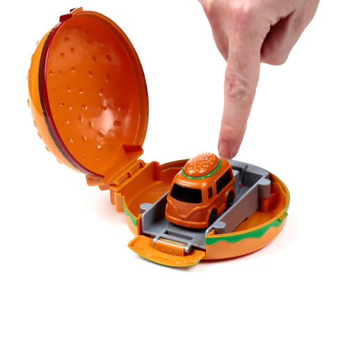 Angled view of a press and launch hamburger truck action toy. The orange burger-shaped case is open, revealing a small orange van with a burger bun roof on a grey launch platform. A finger is poised to press the small vehicle.