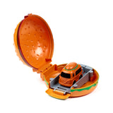Angled view of an open orange press and launch hamburger truck action toy, with its top bun hinged upwards. Inside, a smaller orange toy truck, also with a bun-shaped top, rests on a grey launch ramp. The main toy features a green base.