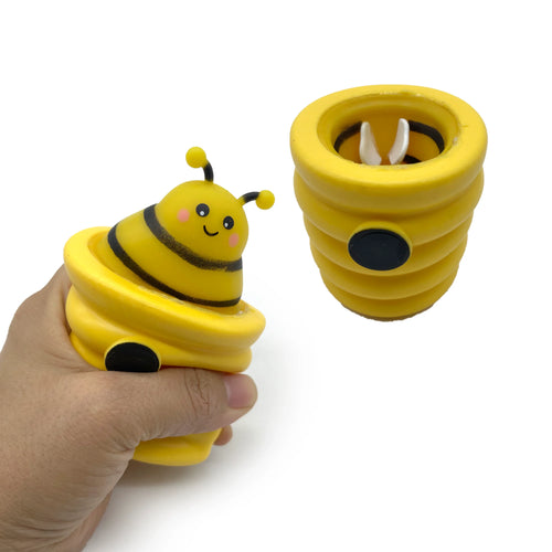 An angled view of a hand squeezing a yellow pop out bee toy, revealing a smiling bee with black stripes emerging from a ribbed beehive. Another yellow beehive with a partially hidden bee is shown beside it on a white background.