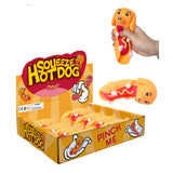 Angled view of an orange display box, labelled SQUEEZE HOT DOG, filled with multiple orange sausage dog fidget toys. One stretchy toy is held by a hand, squeezed to reveal its red 'sausage' filling with yellow 'mustard' detail. Another identical toy lies flat, showcasing its side profile and small pink paw prints.