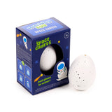 Angled view of a 'Space Cadets' hatching egg toy. The blue box with astronaut graphics and '3+' age rating features a cutout revealing a speckled white egg, with another matching speckled white egg standing alongside.