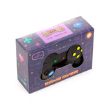 Angled view of a black stretchy game controller fidget toy displayed within its dark purple box. The box features pixelated patterns and text, including 'STRETCHIES' and 'GAME OVER', with a window revealing the controller's yellow, blue, green, and red buttons.