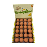 Overhead view of an open display box filled with numerous brown bouncy eggs. The box lid features 'Springtime' text, a bunny, chick, and flowers, with yellow and white checkered sides.