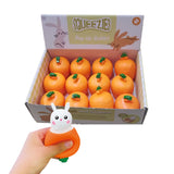 Angled, elevated view of an open display box containing twelve Springtime Rabbit & Carrot Pop Out Toys, each an orange carrot shape with green details. In the foreground, a hand demonstrates one toy, squeezing it to reveal a white rabbit head with black eyes and pink cheeks emerging from the orange carrot.