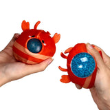 In-context shot of a pair of red lobster fidget toys held by hands. The left toy has a dark blue squishy eye, and the right toy features a clear blue bead-filled eye.