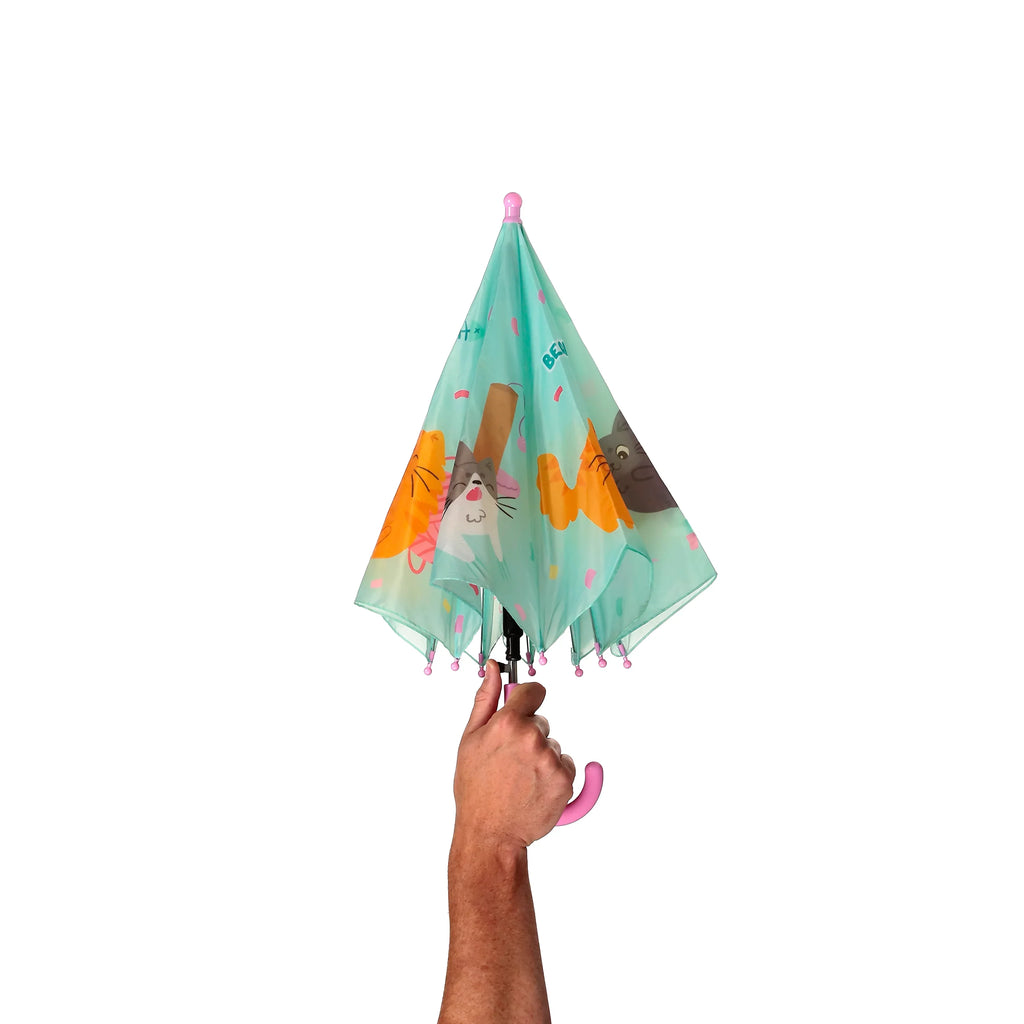 A hand holding a closed kids' umbrella, featuring a turquoise canopy decorated with colourful cartoon cats, pink bobble finials, and a pink curved handle.