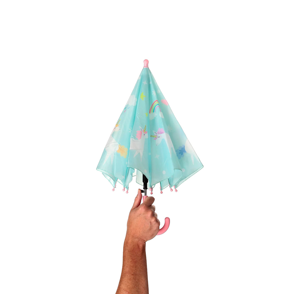 A hand holds a gathered kids' umbrella, its light blue canopy adorned with colourful unicorn, rainbow, star, and cloud patterns. The umbrella features a pink handle and matching pink tips.