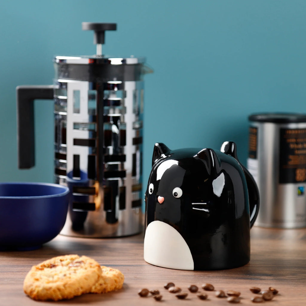 Lifestyle shot of a black ceramic Feline Fine cat-shaped upside down mug, featuring white eyes, a pink nose, and a white belly patch. It is placed on a wooden surface with two biscuits and scattered coffee beans in the foreground, and a chrome cafetière and blue bowl visible in the background against a teal wall.