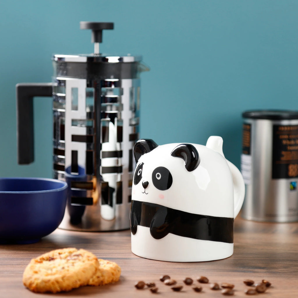 Pandarama Upside Down Ceramic Shaped Mug, featuring a cute white and black panda face, ears, and handle from an angled view in a lifestyle setting on a wooden table with biscuits and scattered coffee beans.