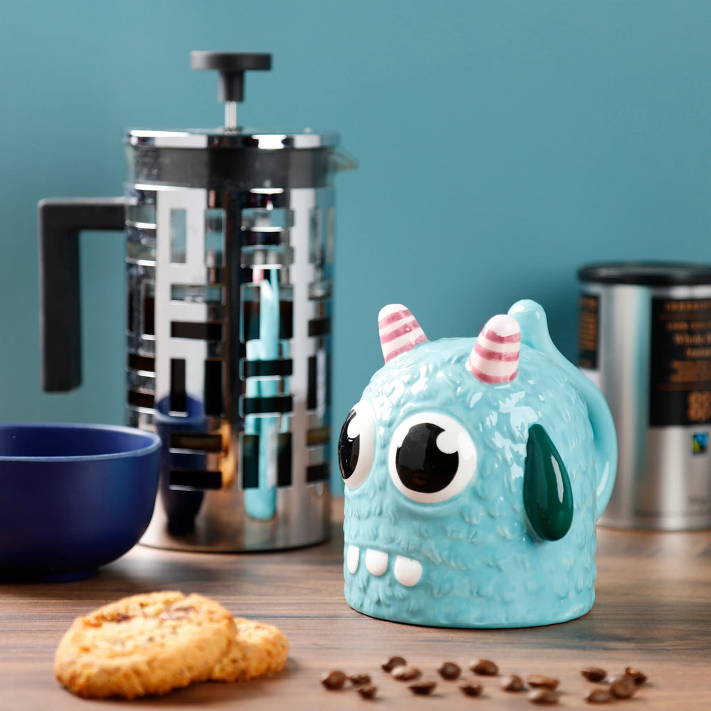 Lifestyle shot showing an angled view of a blue ceramic upside down monster shaped mug, with large black and white eyes, white teeth, and pink and white striped horns. It sits on a wooden table with biscuits, scattered coffee beans, and a cafetière in the background.