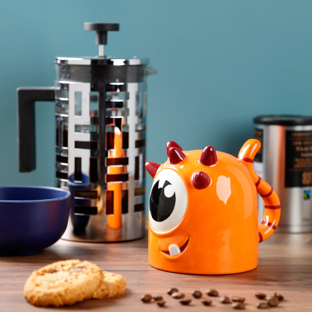Lifestyle shot of an orange monster-shaped ceramic mug, an upside down design with a large eye, red horns, a striped handle, and a single tooth, sitting on a wooden table with a French press, coffee beans, and biscuits.