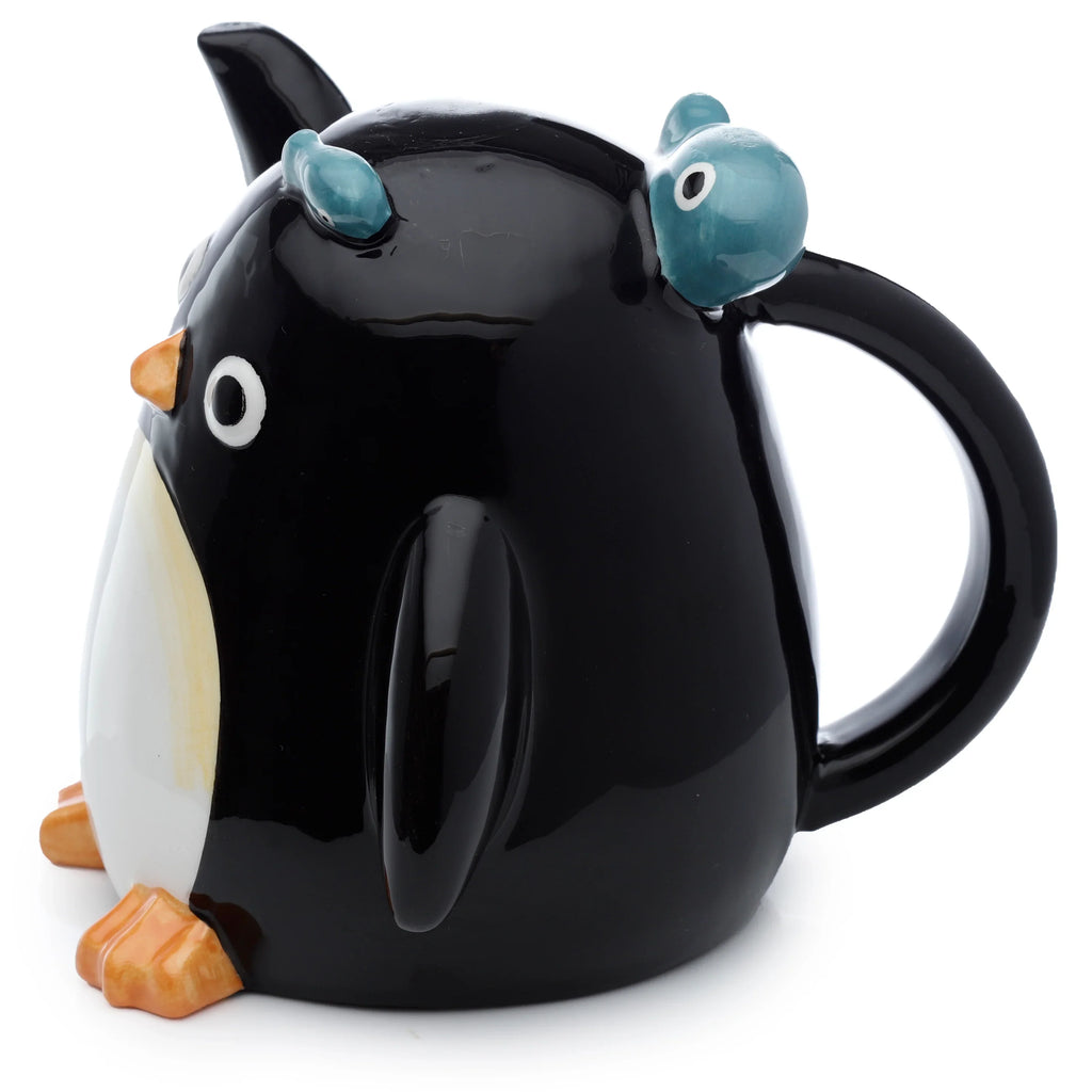 Angled view of a Huddle Penguin Upside Down ceramic shaped mug, featuring a black and white penguin design with an orange beak, orange feet, and two blue baby penguins nestled on its back, complete with a black handle.