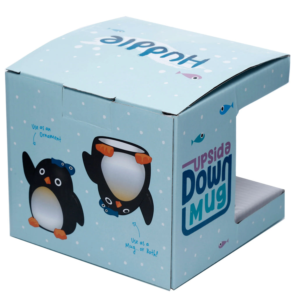 Angled view of a light blue Huddle Penguin Upside Down Mug packaging box, featuring cartoon penguin illustrations and text like 'Use as an Ornament' and 'Use as a Mug... or Both!'. The box has white polka dots, small fish, and is partially open to show a white protective insert.