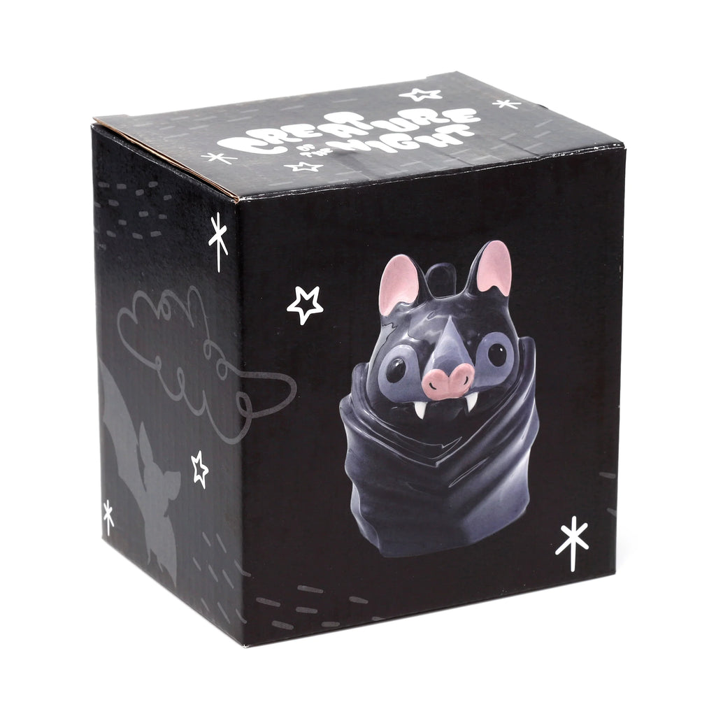 Angled view of a black novelty Upside Down Bat Mug gift box, showing a cartoon bat design with pink ears, nose, and white fangs, plus 'Creature of the Night' text and white star motifs.
