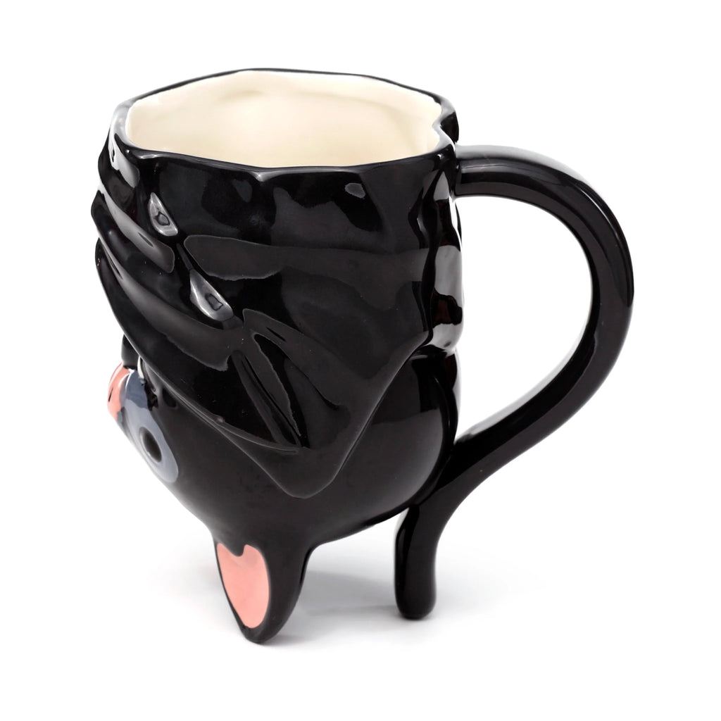 Angled view of a glossy black novelty upside down ceramic bat mug, featuring sculpted bat wings, a handle, visible grey eye, pink nose, and pinkish feet acting as a base, with a cream-coloured interior, on a white background.