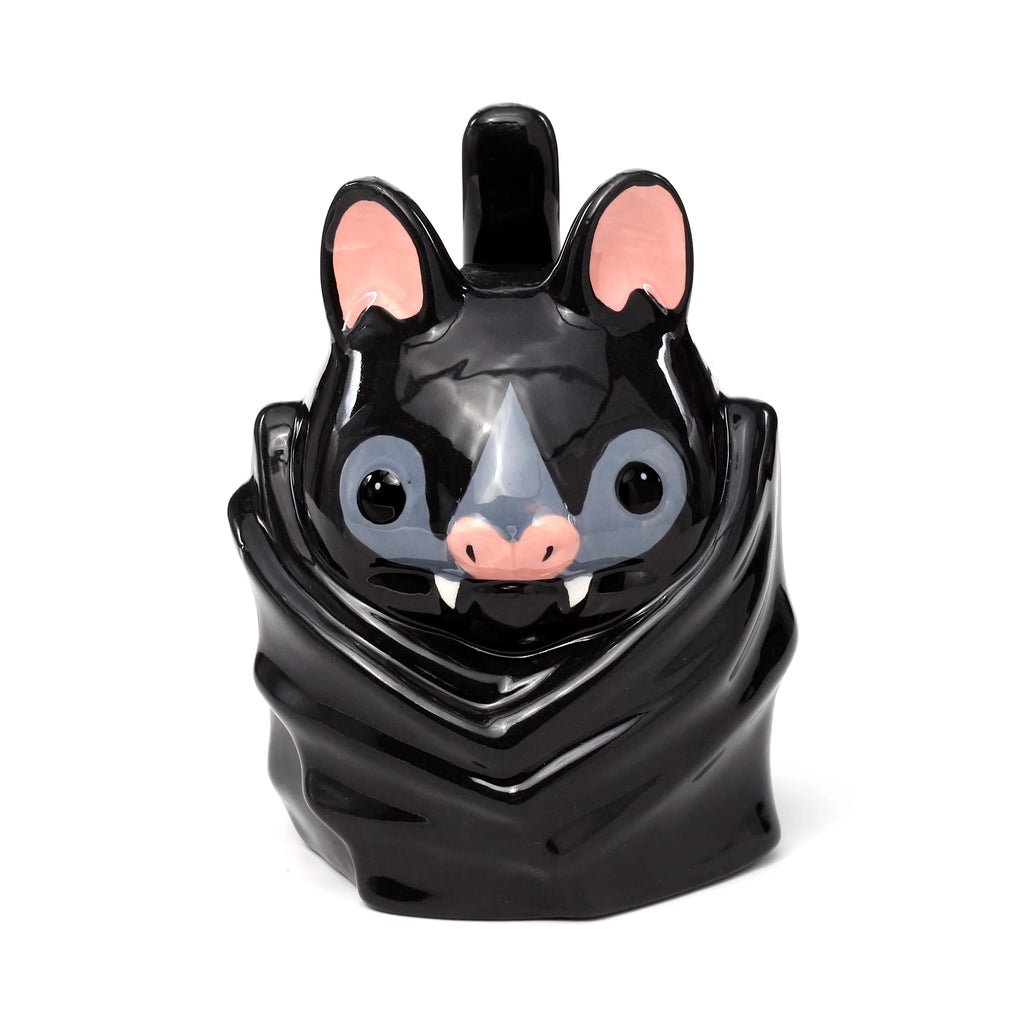 Front view of a glossy black ceramic novelty bat mug, designed to resemble an upside-down bat. It features large pink-lined ears, grey eye patches, a pink snout, and white fangs. A small handle is visible at the top, between the ears.