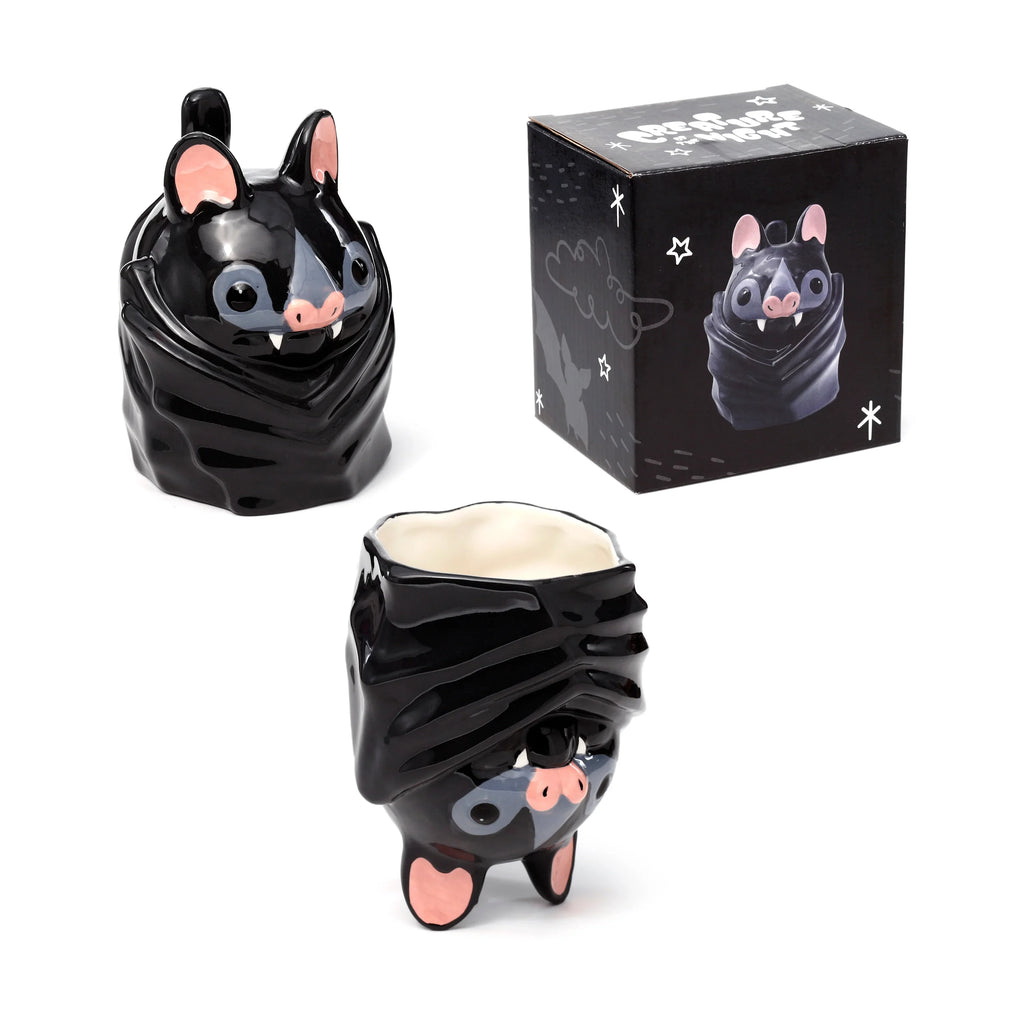 Novelty black ceramic bat mug displayed in two orientations next to its black packaging box on a white background. One glossy mug shows the bat's sleeping face with pink ears, pink nose, white fangs, and grey eye detail, forming the top of the 'upside down' mug, standing on three small pink feet. The second mug is pictured upright, revealing its creamy white interior and the bat's face sculpted into its base. The accompanying black box features white illustrations of bats, stars, and the mug itself.