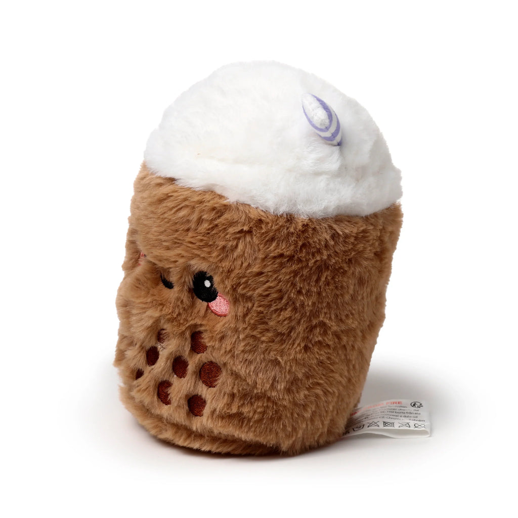 Angled front view of a fluffy, brown and white plush item designed to look like a bubble tea. It features an embroidered winking face, dark brown embroidered tapioca pearl details, a fluffy white top, and a small purple striped straw. A white fabric care label is visible at the bottom.