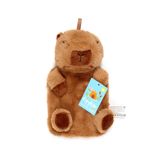 Front view of a brown, fluffy capybara plush hot water bottle cover with stitched eyes, nose, and four small paws. A light blue product tag showing a capybara illustration and 'Capybara Hot Water Bottle' text is attached.