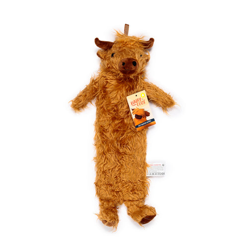 Full-length view of a long hot water bottle with a plush, shaggy ginger-brown Highland coo cover, featuring small horns, a dark snout, and short legs, laid flat on a white background. A product information tag is visible on its side.