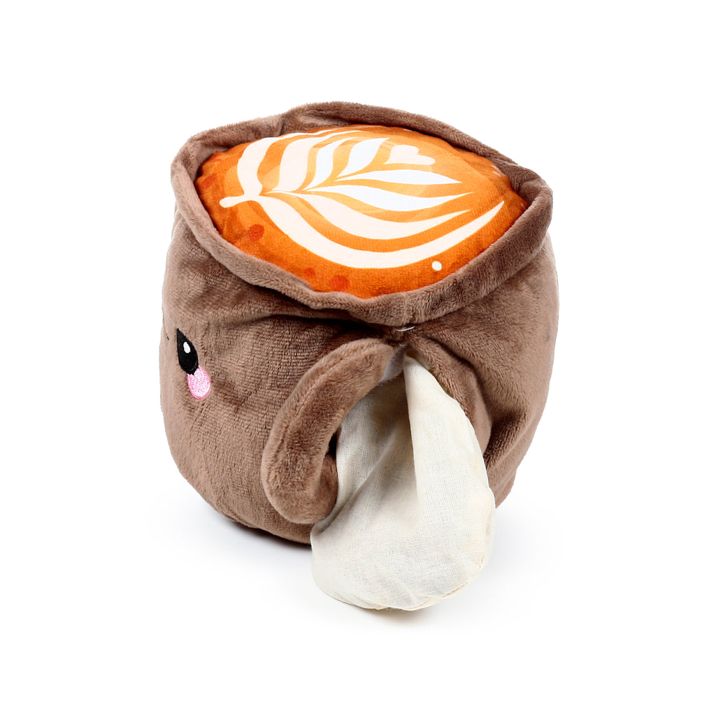 Angled view of a plush brown cappuccino cup-shaped heat pack with an embroidered face, a cream fabric pouch visible on the side, and a cream latte art leaf design on its orange top.